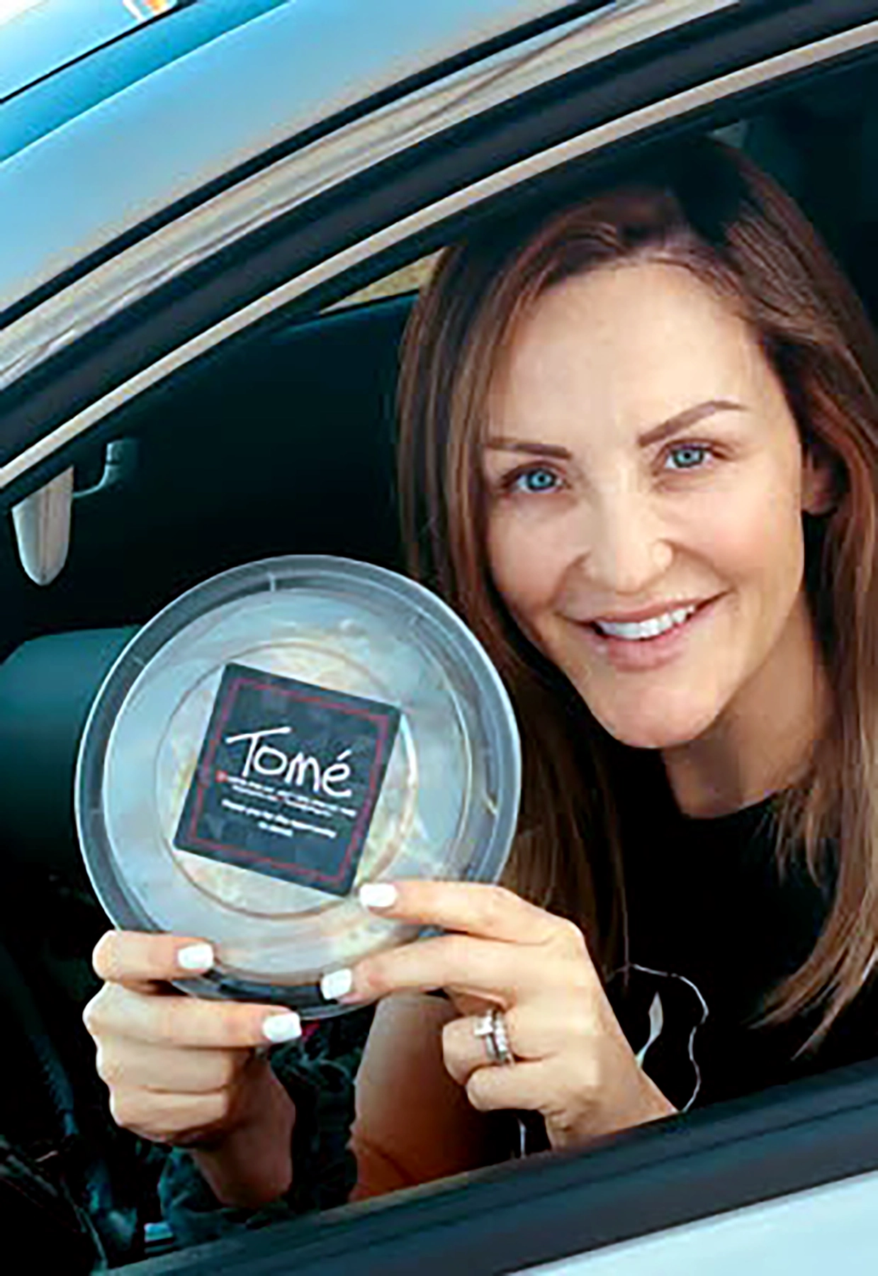 Person in car holding a packaged meal to-go from Tomé, smiling through the drive-thru window for convenient carry-out
