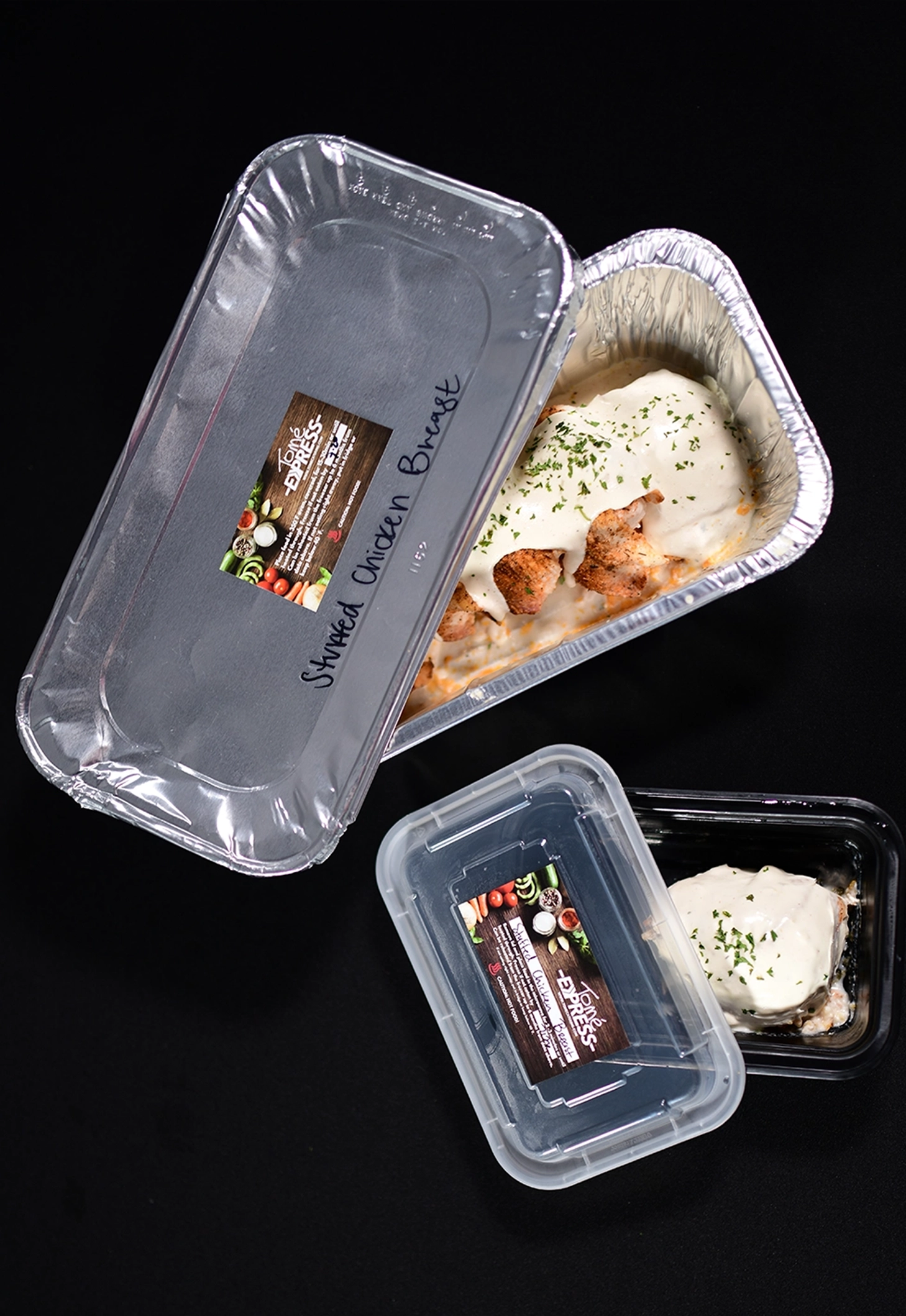 Family style carry-out meals in foil and plastic containers ready for drive-thru pickup, including seasoned chicken breast with sauce